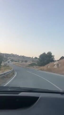 The video is a first-person perspective from inside a vehicle traveling on a paved road. The road curves to the left and continues towards a slightly hilly landscape with sparse vegetation, consisting mostly of dry grass and scattered trees. The sky is clear and pale blue, suggesting it is either early morning or late afternoon. In the distance, a yellow truck with a blue stripe on its roof is visible driving on the same road. The vehicle is equipped with a dashboard and windshield wipers are visible in the foreground. The road has white lines marking the lanes and a metal guardrail on the left side. The terrain appears arid.
