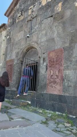 The video shows a medium shot of the entrance to an old stone building, possibly a church. A person wearing a dark blue jacket, dark pants, and a bright blue shirt is seen from behind, stepping through a metal grate that bars the entrance. Above the entrance, a security camera is mounted on the stone wall. On either side of the entrance, large carved stones with intricate cross designs are embedded in the wall. The building's facade is made of rough, dark stone blocks, some of which appear weathered and discolored. The sky is clear and blue, indicating fair weather. To the right of the building, a pathway leads to a railing overlooking a body of water, suggesting the building is situated in a scenic location.
