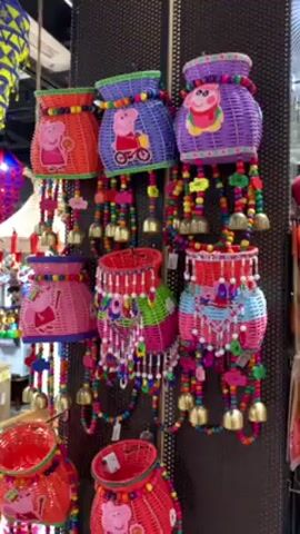 The video shows a shop selling various colorful decorative items. The focus is on a display of hanging decorations featuring woven baskets adorned with Peppa Pig images, colorful beads, and small bells. These decorations are in multiple colors, including orange, blue, purple, pink, red, and green. The background reveals other items for sale, such as geometric patterned hanging lamps, hats, and various small objects on shelves. The shop appears to be well-lit and filled with a diverse selection of merchandise.