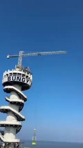 The video shows a person zip-lining from a tall tower with the word “BUNGY” in large letters on it. The tower also has a spiral staircase. A crane stands atop the tower, from which the zip line extends. The person is seen hanging from the zip line against a clear blue sky. A small, green and yellow cage-like structure hangs suspended from a cable below the tower. The environment appears to be near a body of water, visible in the background.