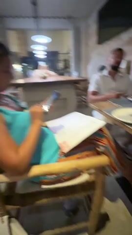 The video opens with a woman in a blue dress sitting at a wooden table, holding a mobile phone and looking at a menu. She is seated on a chair with a colorful blanket draped over it. To her right, a man in a white shirt is gesturing with his hands while looking at the camera. Further down the table, another woman in a white top is using her phone. Next to her, a man in a black t-shirt is looking at a menu. Several menus, plates, cutlery, water bottles, and glasses are on the table. The environment appears to be a restaurant with stone walls and modern light fixtures. The scene is captured from a handheld perspective, with some motion blur.