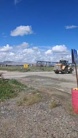A construction site under a bright blue sky with scattered clouds. A yellow bulldozer is visible in the middle ground moving across a dirt road. In the background, a partially constructed metal building stands, and further in the distance, a large pile of debris or earth is present. A yellow sign is posted on the left side of the road. The foreground consists of gravel and grass.