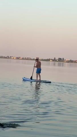 The video shows two men on paddleboards on a calm body of water. In the background, there is a distant shoreline with some buildings and trees visible. The sky is a pale pinkish-orange, suggesting it is either sunrise or sunset. The man in the foreground is closer to the camera, seated on his paddleboard and holding a paddle. The man in the background is standing on his paddleboard and paddling. The water is slightly rippled, and reflections of the sky and the paddleboarders are visible on the surface. The overall atmosphere appears peaceful and recreational.