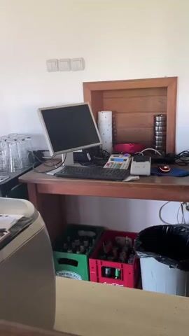 The video shows a cluttered workspace with an old computer, a cash register, and various bottles of liquor in a wooden cabinet.  There is also a trash can and crates of soda and beer under the desk.