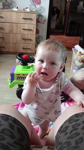 A baby is standing in front of the camera, holding a finger to their mouth. The baby is wearing a white onesie with a pattern of pink hearts and leaves. The baby is looking at the camera with a curious expression. Behind the baby is a green toy tractor with a red steering wheel. In the background, there is a wooden dresser and a doorway leading to another room. The floor is covered with a patterned rug and a pink blanket. The image is taken from a low angle, as if the camera person is also sitting on the floor.