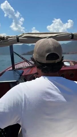 This is a POV shot from behind a person wearing a white t-shirt and a tan baseball cap, sitting in the driver's seat of a red boat. The person is facing away from the camera, steering the boat. The boat has a tan canopy overhead and a wooden dashboard with instruments. The background shows a body of water with mountains and a town on the shore under a bright blue sky with scattered white clouds. The boat appears to be moving through the water, indicated by the slight blur of the scenery. The overall scene depicts a leisurely boat ride on a sunny day.