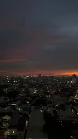 A high-angle aerial shot captures a cityscape at dusk. The sky is a deep, bruised purple, transitioning to a fiery orange glow on the horizon where the sun has set. Wispy clouds are scattered across the sky, tinged with pink and red from the fading light. Below, the city is a dense tapestry of buildings, a mix of low-rise structures and taller buildings interspersed throughout. Streetlights and illuminated windows twinkle like scattered jewels, creating a warm, inviting contrast to the darkening sky. The overall impression is one of a city settling into the night, bathed in the last vestiges of daylight.