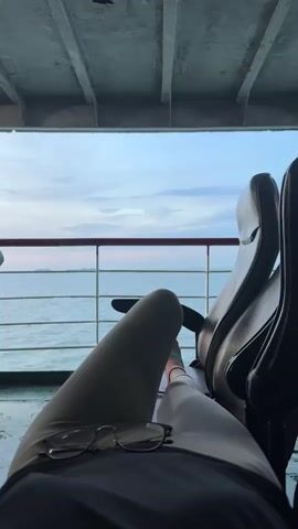 The video shows a first-person perspective of someone relaxing on a ferry. The camera is pointed downwards, focusing on the person's legs, which are extended in front of them, and a pair of glasses resting on their lap. The person is wearing light-colored leggings. In the background, there is a large window offering a view of the ocean and the sky. The sky appears overcast with some hints of pink from a sunset or sunrise. The ocean is calm, and a railing is visible outside the window. Two empty seats with headrests are positioned to the right of the frame, suggesting other passengers might be present or have been there. The ceiling of the ferry, with visible beams and structures, is also in the frame, indicating the indoor seating area. The floor appears to be a green, slightly wet surface. The overall impression is one of peaceful travel.