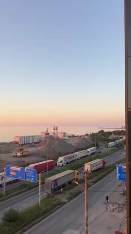 The video pans across a highway at sunset. A long line of semi-trailer trucks is backed up on the highway, and several more are parked on the side. In the background, there is an industrial area with large piles of gravel and several buildings, possibly a cement plant. The sea is visible beyond the industrial area, with a ship on the horizon. The sky is a gradient of orange, pink, and blue. There are a few people visible walking on the side of the road. The overall scene depicts a transportation bottleneck in an industrial coastal area during sunset.