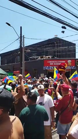 The video shows a large crowd of people celebrating in the street. Many are waving Venezuelan flags, wearing Venezuelan-themed clothing, and carrying other paraphernalia such as a large red horn being played. The crowd appears to be on a city street lined with apartment buildings and commercial properties. Shoes are hanging from powerlines overhead. The sky is blue and clear, suggesting a sunny day.