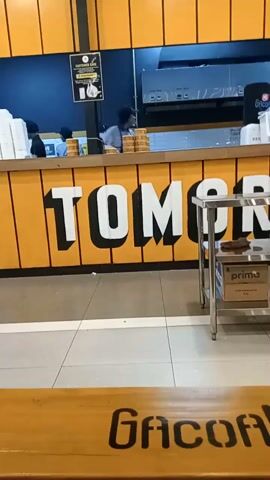 The camera pans up to show a restaurant counter with a yellow front panel and white lettering that says "ITS TOMORROW". Behind the counter, there are three workers wearing light blue shirts and face masks, assembling orders. Stacks of plates and food containers are visible on the counter. The area above the counter is dark blue, and a sign labeled "CUSTOMER CARE" is posted. A stainless steel cart holding a brown shoe and a "Prima" box is located to the right of the counter. The floor is tiled, with a yellow section in the lower part of the frame bearing the logo "GACOAN".