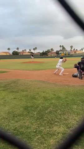 A baseball game is in progress. A batter, wearing a white uniform with the number 22, swings at a pitch. The ball goes into the field. An umpire is on the field. The catcher is wearing protective gear. Other players are visible. The field is green grass and brown dirt. There is a black chain-link fence partially obscuring the foreground. There are houses and palm trees in the background, under a gray sky. The scoreboard is visible.