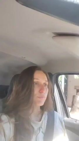 Here's a description of the video:

A woman with long brown hair and wearing a light-colored t-shirt is seated in the driver's seat of a car. She is wearing a seatbelt. The car appears to be moving, with buildings visible outside the window. The interior of the car is visible, including the ceiling and sun visor. The woman looks towards the side and then directly at the camera, then the camera is angled upward towards the car's ceiling. There is music playing.