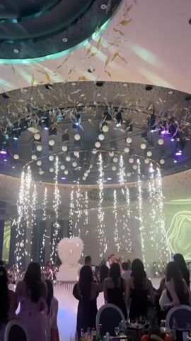 The video shows a large room, probably a wedding hall, with fireworks falling from a circular lighting fixture hanging from the ceiling. A large white flower-shaped cake is in the background, partially obscured by smoke. People are standing around, some holding up their phones to record. The lighting fixture has many white orbs and black spotlights. Above the fixture, there is another decorative structure with many shiny, abstract shapes resembling leaves or birds. The room is filled with tables and people in formal attire, suggesting a celebration or formal event.