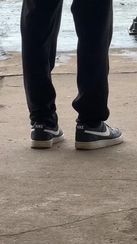 The video shows a close-up, low-angle view of a person's lower legs and feet. The person is wearing black sweatpants and black Nike sneakers with white accents. The sneakers have the Nike logo and the word "NIKE" on the heel. The person is standing on a concrete surface, which appears to be outdoors and somewhat damp. The background is blurred, but suggests an industrial or unkempt outdoor environment. The footage is static, with no movement from the subject.