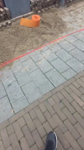 The video shows a street construction scene in front of a store. There are construction workers present, wearing orange vests, working on what appears to be a sidewalk or pavement. They are laying down bricks and possibly compacting the ground with a vibratory plate compactor. Some materials, including sand and bricks, are visible at the construction site. A red cable or hose runs along the ground, delineating the work area. The store windows display mannequins and clothing. The overall environment suggests an urban setting during daytime.