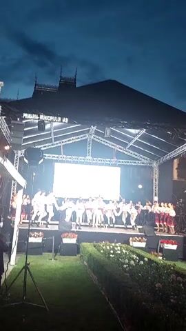 This video captures an outdoor performance on a stage at dusk. A group of young people, dressed in traditional Romanian folk costumes, are dancing in front of a large LED screen. The costumes appear to be white with red and black embroidery, and the boys are wearing hats. They are holding hands in a circle as they dance. Behind them, a band is visible, also in traditional attire, playing instruments.

The stage is set up with lighting rigs and scaffolding. The LED screen displays a graphic with intricate patterns, possibly a cultural motif, and text. The visible text on the screen reads "Ansamblul Folcloric de Copii si Tineret" and "Florile Moldovei," along with "Iasi." Above the stage, on the facade of a dark building, text reads "PRIMARIA MUNICIPIULUI IASI," indicating the municipality of Iași.

The sky is dark blue, suggesting it is either early evening or late dusk. The lighting on the stage is bright, illuminating the performers and the screen. In the foreground, there is a grassy area with planters containing flowers, some of which are white and red. The overall atmosphere appears to be festive and celebratory, likely a cultural event or festival taking place in Iași, Romania.