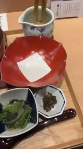 The video shows a Japanese meal set arranged on a wooden tray. The main dish appears to be Oden, served in a metal pot. It contains ingredients like eggs, fish cakes, and other ingredients in broth.  Next to the Oden is a red bowl with a white piece of paper inside, and a small blue and white dish containing vegetables, possibly some type of salad or pickled greens.  There is also a tiny dish containing what looks like pickles. A small bowl of cooked rice sits on the tray as well. The setting seems to be in a restaurant.