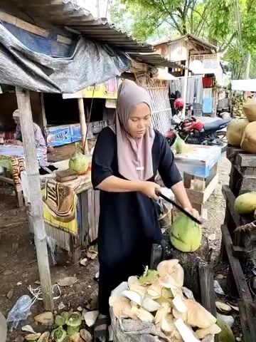 A person wearing a hijab and a black dress is cutting a green coconut with a large knife or machete. The person is standing at a small wooden stand that is cluttered with other coconuts. The coconut being cut sits on a tree stump. There are other coconuts on shelves to the right. The background shows a roadside scene with other small shops and a motorcycle parked nearby. The area appears to be a rural or semi-rural location, possibly a market.