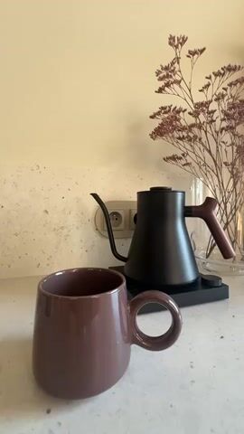 A hand is shown picking up a black gooseneck kettle from its base. The kettle is then tilted and water is poured from it into a brown mug. The mug is on a countertop in front of a speckled backsplash. A vase with dried flowers is to the right of the kettle. An electrical outlet is visible behind the kettle.