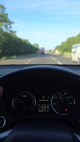 The video shows a first-person view from inside a car driving on a highway. The driver's hands are not visible, but the steering wheel and dashboard are in focus. The dashboard displays a low tire pressure warning light, a speedometer reading approximately 60 mph, an odometer reading 003047 miles, and an exterior temperature of 17 degrees Celsius. The road ahead is clear, with other vehicles visible in the distance, including a truck and a white van. Trees and vegetation line both sides of the highway, and the sky is blue with some clouds. The overall impression is of a normal drive on a sunny day, with the exception of the low tire pressure warning.