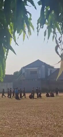The video shows a group of young people on a dry, dusty field. They appear to be engaged in some kind of outdoor activity or practice. There are several individuals standing and a few sitting. In the background, a building with a distinctive roof shape is visible, along with some fencing and foliage that frames the top of the shot. The lighting suggests it is daytime, possibly late morning or afternoon.