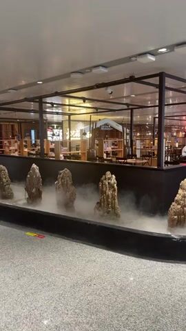 The video shows a man in a white shirt and black pants working behind a counter in a restaurant. The restaurant appears to be open and customers are seated at tables in the background. The foreground of the video features a decorative water feature with rocks and mist. The overall environment is an indoor restaurant setting with modern decor.