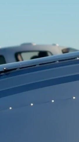 This is a close-up, side-view shot of a white, single-engine aircraft in flight. The cockpit is visible, showing two people wearing headsets and appearing to be piloting the plane. The right wing and landing gear, which consists of two streamlined pods, are prominently displayed. The background is a clear, pale blue sky, indicating daytime. The plane appears to be in the process of takeoff or flight.