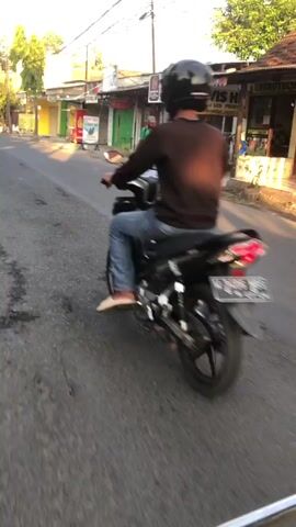 The video shows a view from a motorcycle traveling on a street in what appears to be a village or small town. The rider is wearing a black shirt and a helmet, and is moving forward. Another motorcycle is visible ahead of the rider, also moving in the same direction. The street is lined with buildings on the right side, which appear to be shops or businesses. One of these buildings has a sign that reads "FOTOCOPY" and another has a sign advertising "SERVIS". A person wearing a green shirt and a sarong is walking on the sidewalk. There are parked motorcycles in front of the buildings. The sun is shining, casting shadows, suggesting it is daytime, possibly late afternoon. The road surface is asphalt with some visible wear and tear. The overall environment seems to be a typical street scene in Southeast Asia.