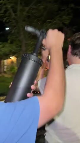 A video shows four men around a fire pit at night. The man in the foreground, wearing a blue t-shirt, is holding a black pump. He aims the pump towards the fire pit. The man next to him, wearing a white t-shirt, is holding a long, black snake-like object. He is spinning the object above his head. Another man, wearing a black t-shirt and shorts, is standing in the background. He is holding a drink in his hand. The fourth man is partially visible behind the man in the blue shirt. The fire pit is in the center of the frame, with flames reaching up. There is a green can and a box of snacks on a wooden table next to the fire pit. Trees and bushes are visible in the background.
