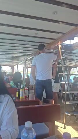 The video pans across the interior of a boat, showing a seated area with wooden tables and benches. Several people are visible, some seated and others standing. In the foreground, a person in a white t-shirt and denim shorts stands near a ladder leading to an upper deck. On a table, there are several bottles, possibly drinks. The boat appears to be on the water, with a coastline and buildings visible in the background. The overall impression is a casual setting on a boat trip.