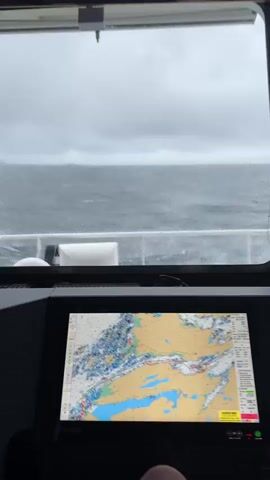 The video shows the view from the bridge of a vessel, looking out a window and at a navigation screen. The window is wet with rain, and a windshield wiper is in operation, clearing the view of the choppy, gray sea and overcast sky. The navigation screen displays a nautical chart with depth soundings, buoys, and landmasses. The vessel appears to be in motion.