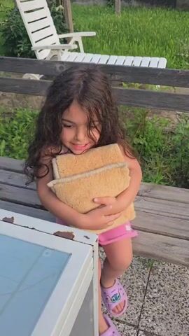 A young girl with long, dark, curly hair is holding a tan-colored object with a textured surface in her arms. She is wearing a white tank top with colorful stripes and pink shorts. She is also wearing pink sandals with a purple cartoon character on them. The girl is sitting on a wooden bench, and she is looking to her right with a smile. In the background, there is a white lawn chair and a wooden fence. The setting appears to be outdoors, possibly in a backyard, with green grass and foliage visible.