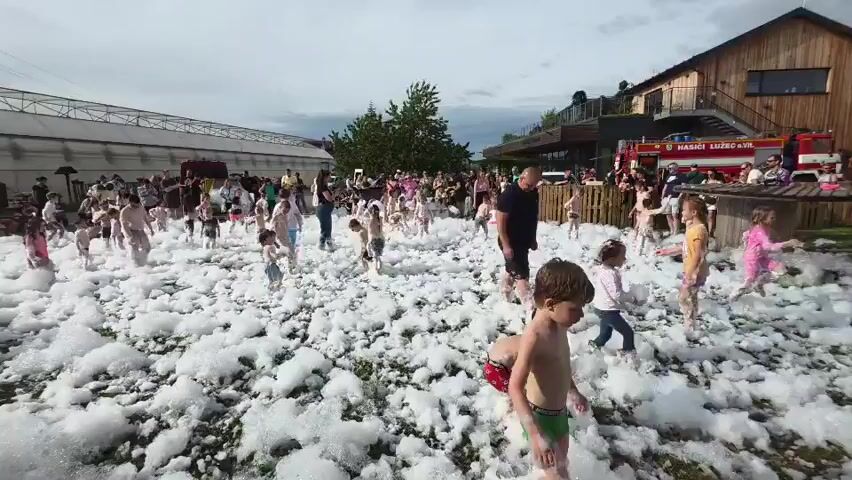A wide shot captures a children's event on a grassy field covered in thick, white foam. Many children are playing and running through the foam, some are partially covered, and others are nearly submerged. 
Adults, mostly standing on the edge, watch the children. In the background, there's a crowd of people near a fire truck with the words "HASIČI LUŽEC AVL" visible. Behind the crowd is a modern wooden building with a deck and stairs. To the left of the frame is a large greenhouse-like structure. The sky is cloudy and overcast.