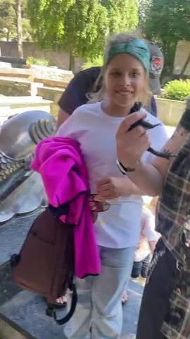 A person is holding a toad. The toad is brown and has bumpy skin. The person's hand is around the toad's body. Other people are in the background. One person is wearing a pink jacket and another is wearing a blue denim jacket. There are trees and a stone structure in the background. The sun is shining.
