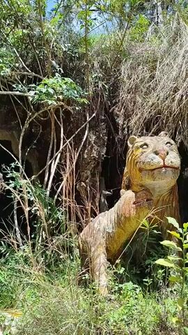 A person with a yellow hard hat and a black backpack walks into a small, dark cave opening in a jungle environment. The cave opening is surrounded by green foliage, trees, and rocks. A dinosaur statue is partially visible on the right side of the frame.