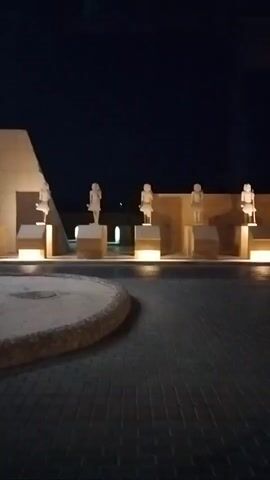 The video shows a row of six white statues on pedestals at night. The statues appear to be of people and are lit from below. The background behind the statues is a dark wall. In the foreground is a paved area. The video is taken from a moving vehicle, as the perspective changes slightly. The lighting is low, emphasizing the illuminated statues.