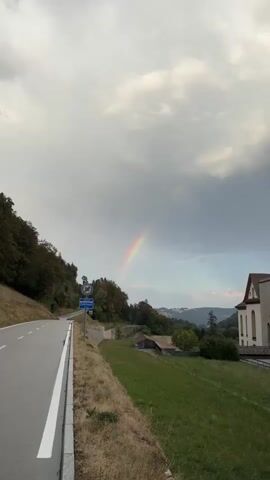 The video shows a winding road with a white line marking the shoulder. On the left side of the road, there are trees and a slope. On the right side, there is a grassy area, some houses, and a large building with a white facade and a reddish roof. In the distance, there are rolling hills. A rainbow is visible in the cloudy sky. There are also road signs, one of which indicates a speed limit of 50. The weather appears to be overcast with some patches of lighter sky where the rainbow is. The video is shot from a static position, likely handheld.