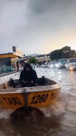 The video depicts a flooded street in Yorkatay, Uzbekistan, during what appears to be daylight hours under an overcast sky. The primary subject is a yellow boat with "TAXI 1260" written on its side, being navigated through the deep water by a person wearing a hooded jacket. The boat is creating a wake as it moves, indicating the presence of significant water depth.

The scene is a city street, with businesses and buildings lining the sides. Cars are visible submerged in the water, some appearing to be stalled or abandoned. The reflections on the water's surface suggest a cloudy or possibly rainy weather condition. The overall atmosphere conveyed is one of disruption and unusual circumstances due to the flooding. There's a sense of ingenuity in using a boat as a mode of transport in such a situation.