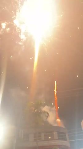 The video shows fireworks exploding in the night sky. In the foreground, there is a bright light source on the left side of the frame, possibly a streetlight or a camera flash. The sky is dark, with some wisps of smoke from the fireworks. There are also some power lines visible in the distance, and what appears to be the roof of a building. The fireworks are colorful, with bursts of pink and orange. One firework is a large, spherical explosion of pink, while another is a trailing streak of orange. The video is shaky, suggesting it was filmed by someone holding a camera by hand.