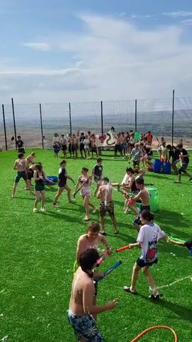 A group of young people are engaged in a water fight on a bright, sunny day. They are on a large, artificial turf field surrounded by a black chain-link fence. In the background, there are hills and a valley. The participants are mostly wearing shorts and t-shirts. Many are holding water guns or buckets, and water is being sprayed in various directions. Some individuals are running, dodging, or taking aim with their water weapons. Several blue and green plastic buckets are scattered across the field. A large banner with a black and white symbol is visible in the background. The ground is wet in many areas, indicating active water play. The overall atmosphere appears to be energetic and playful.