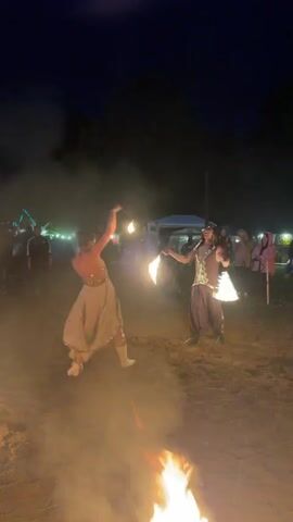 Two performers are engaged in a fire dancing routine at night. On the left, a person in a light-colored, flowing outfit is bending backward, holding a fiery object in their right hand. On the right, a person in darker, ornate clothing is spinning with a fiery object in each hand. The ground appears to be sand or dirt, with a large bonfire in the foreground. In the background, a crowd of spectators is visible, along with some tents and string lights, indicating an outdoor event. The overall atmosphere is dark with the glow of fire and ambient lighting.