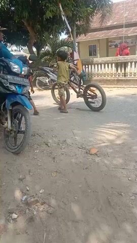 The video opens with a close-up of a blue motorcycle parked on a dirt road. A man wearing a blue shirt and a cap is sitting on the motorcycle, looking to the right. To the right of the motorcycle, a child in a camouflage outfit is standing next to a bicycle with a red frame. The child appears to be interacting with the bicycle. In the background, there is another motorcycle and a building with a white fence. A flag is visible on a pole. The scene suggests an outdoor setting, possibly a village or rural area, with signs of everyday life. The video is shaky and handheld, indicating it was likely recorded by someone walking or moving while filming. The overall impression is one of a casual, candid observation of daily activities.