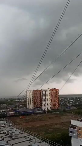 The video captures an elevated view of a town or city, likely Kondratovo, Russia, under a heavily overcast and dark sky, suggesting an approaching or ongoing storm. In the foreground, the top of a building, possibly a rooftop, is visible with gray, flat surfaces. Several dark power lines stretch across the upper portion of the frame from left to right, angled downwards towards the distant landscape.

The middle ground is dominated by two prominent, tall residential buildings, distinguishable by their white and orange-brown facades. These buildings are surrounded by a mix of undeveloped land and other, smaller structures, including what appears to be a commercial building with a blue roof. In the far distance, a hazy curtain of rain is visible against the dark sky, indicating heavy precipitation. The overall landscape shows a mix of urban development and open, possibly rural, areas, with a horizon line of distant houses or buildings. No people or visible text are present in the video.