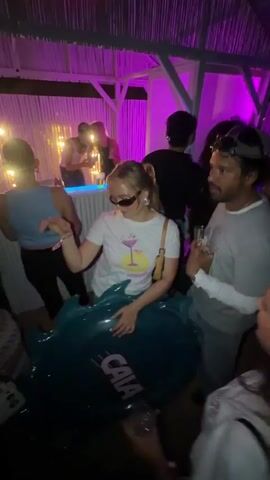 A crowded, dimly lit indoor party with purple lighting features a fair-skinned woman wearing sunglasses, a white graphic t-shirt, and a small handbag, seated on a large, inflatable teal object with "CAIA" printed in white. She is surrounded by several people, including a fair-skinned man in a white long-sleeved shirt and a baseball cap holding a glass with a straw, who turns his head to the left and then to the right while talking to others. In the background, more people are dancing and socializing under yellow stage lights.