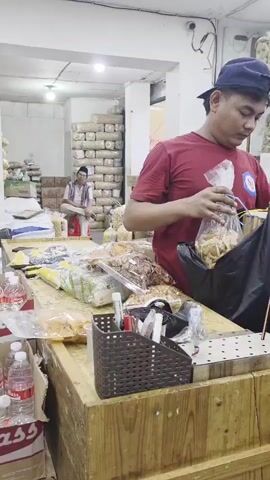 A man wearing a red t-shirt and a blue baseball cap is bagging snacks. He is standing behind a counter filled with bags of snacks. A woman wearing a tan hijab is standing in front of the counter and is holding a black plastic bag with two bags of snacks inside. She is looking at the man. In the background, there are shelves stacked with bags of snacks.