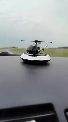 A close-up shot focuses on a miniature helicopter toy mounted on a car's dashboard. The toy helicopter has a black and silver body with spinning blades. The dashboard surface is dark and textured. In the background, a road curves to the left, bordered by green fields under a cloudy sky. Raindrops are visible on the windshield, suggesting it is raining. The camera is positioned at a low angle, looking towards the helicopter.