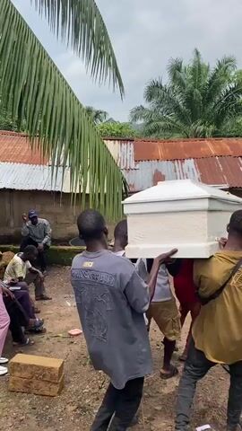 A group of men are carrying a white coffin in an outdoor, rural setting. In the foreground, a man wearing a blue and black shirt is looking away from the camera. Behind him, four men are carrying the coffin, which is held at shoulder height. They are walking away from the camera towards a small, weathered building with a corrugated metal roof. Several other people are visible in the background, seated or standing near the building. Palm fronds and lush green foliage suggest a tropical environment. The ground is a mixture of dirt and scattered rocks. The sky is overcast.