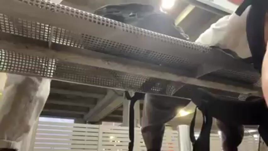 A person is rummaging through luggage placed on an overhead rack, seemingly at a train station or bus terminal. The luggage appears to be in two bags. The individual has brown hair, is wearing a white tank top and gray pants. The rack is made of metal and has a grid-like structure. The environment is a transportation hub with visible concrete structures and a slatted wall. There is ambient light from above.
