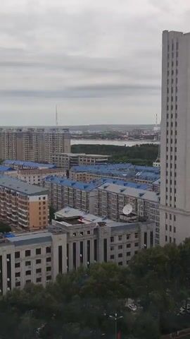 The video shows an aerial view of a city with numerous buildings. The buildings are mostly residential, with many featuring blue roofs. There are also some taller modern buildings in the distance. Trees are visible in the foreground, and a body of water can be seen in the background. The sky is overcast. The video is shot from a high vantage point, possibly a drone or a tall building. There are no people visible in the video.